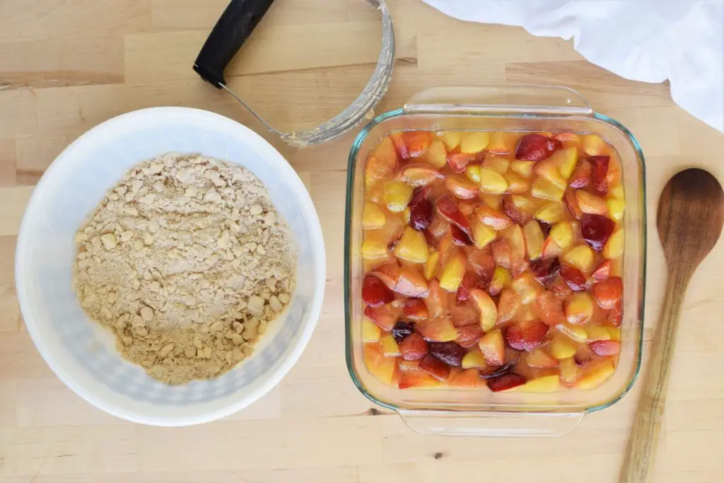 A bowl of dry ingredients with butter cut in next to a 9x9 baking pan with plums.