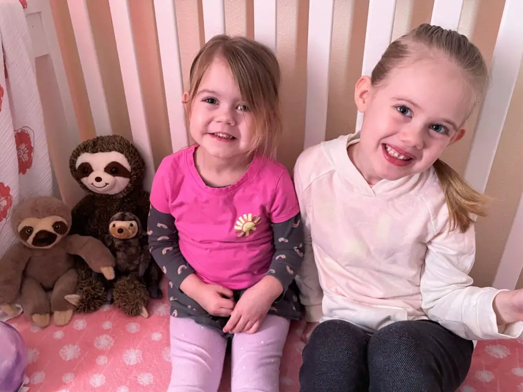 Two girls sitting on a bed with sloth stuffed animals.