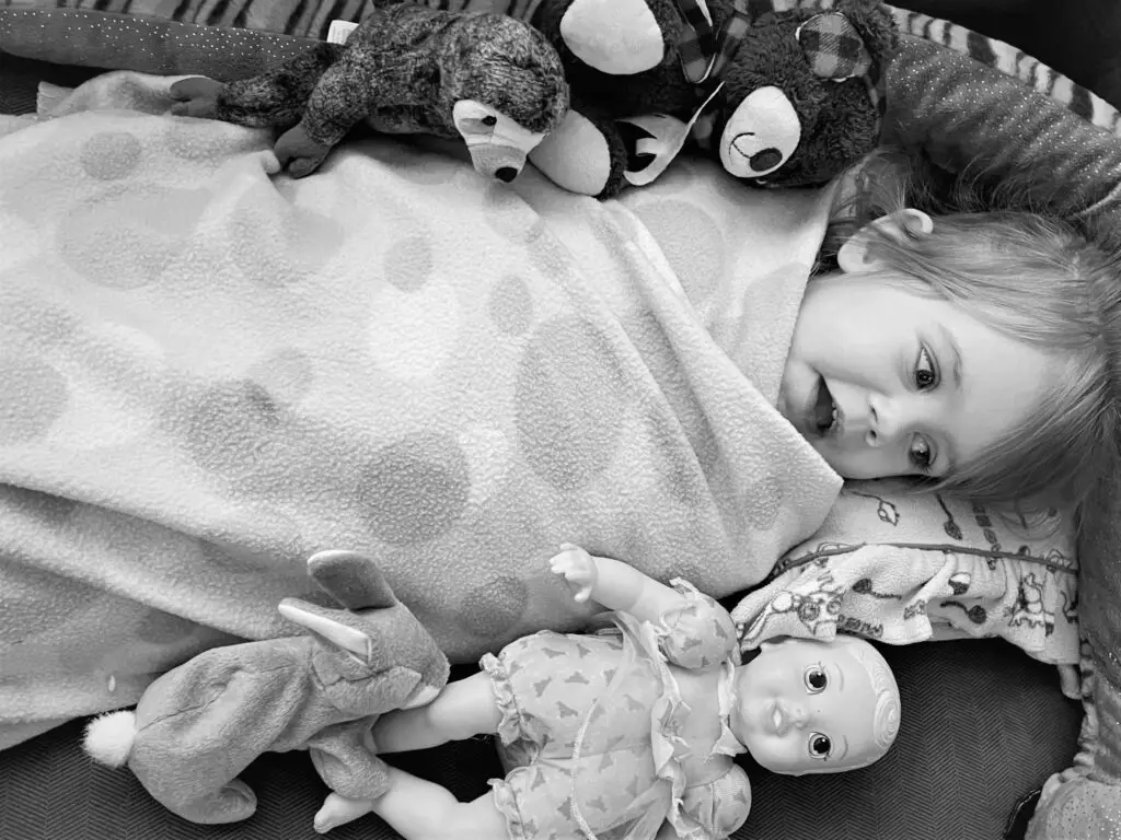 Girl swaddled in blanket surrounded by stuffed animals and a doll.