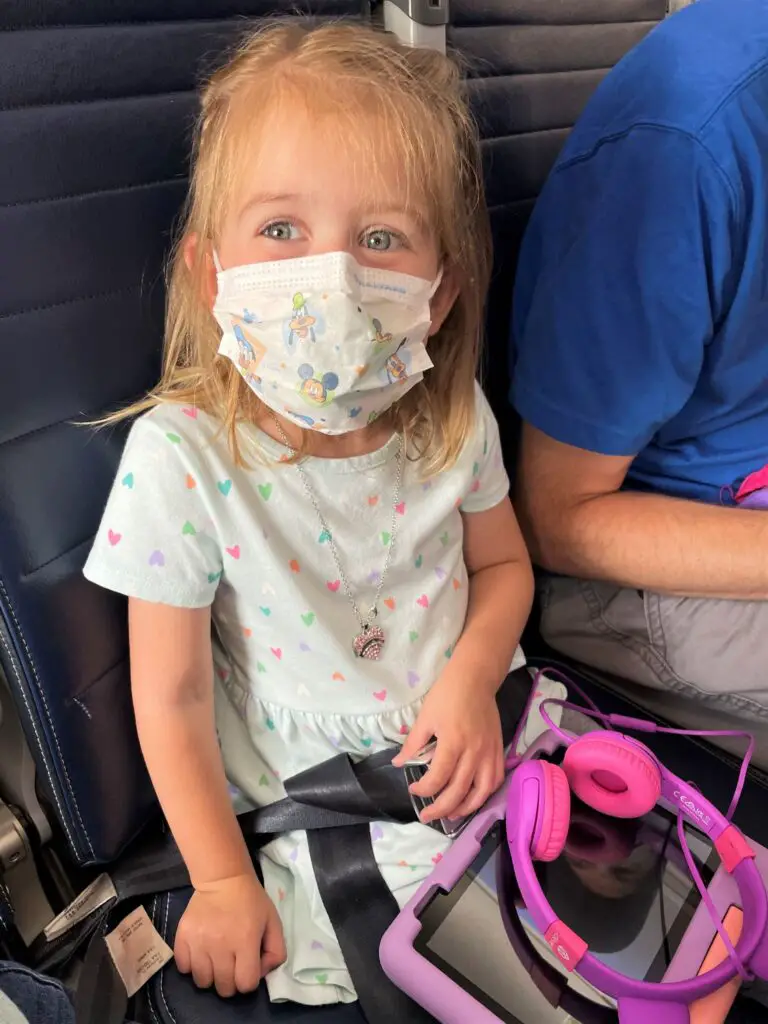 Ellie sitting on the plane with a mask on and purple tablet and headphones.