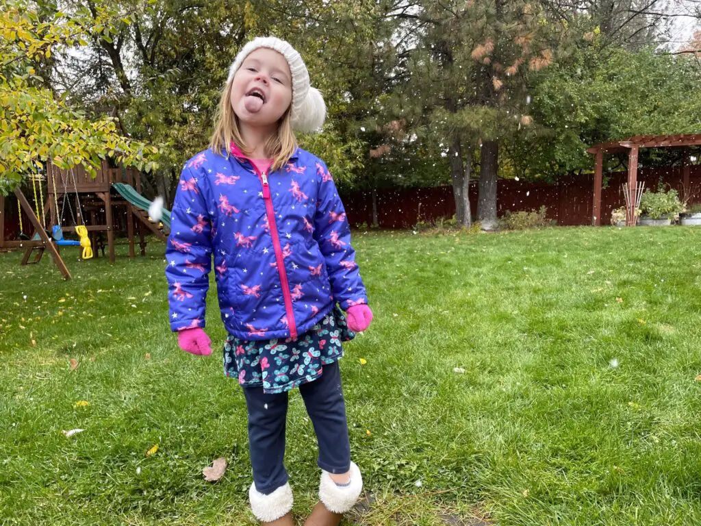 Ellie in blue in pink coat standing in backyard with tongue sticking out. Some snowflakes falling.